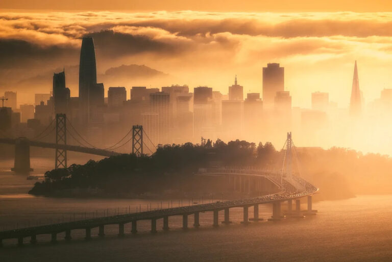 City skyline at sunrise with a curved suspension bridge spanning the water and a hazy orange glow.