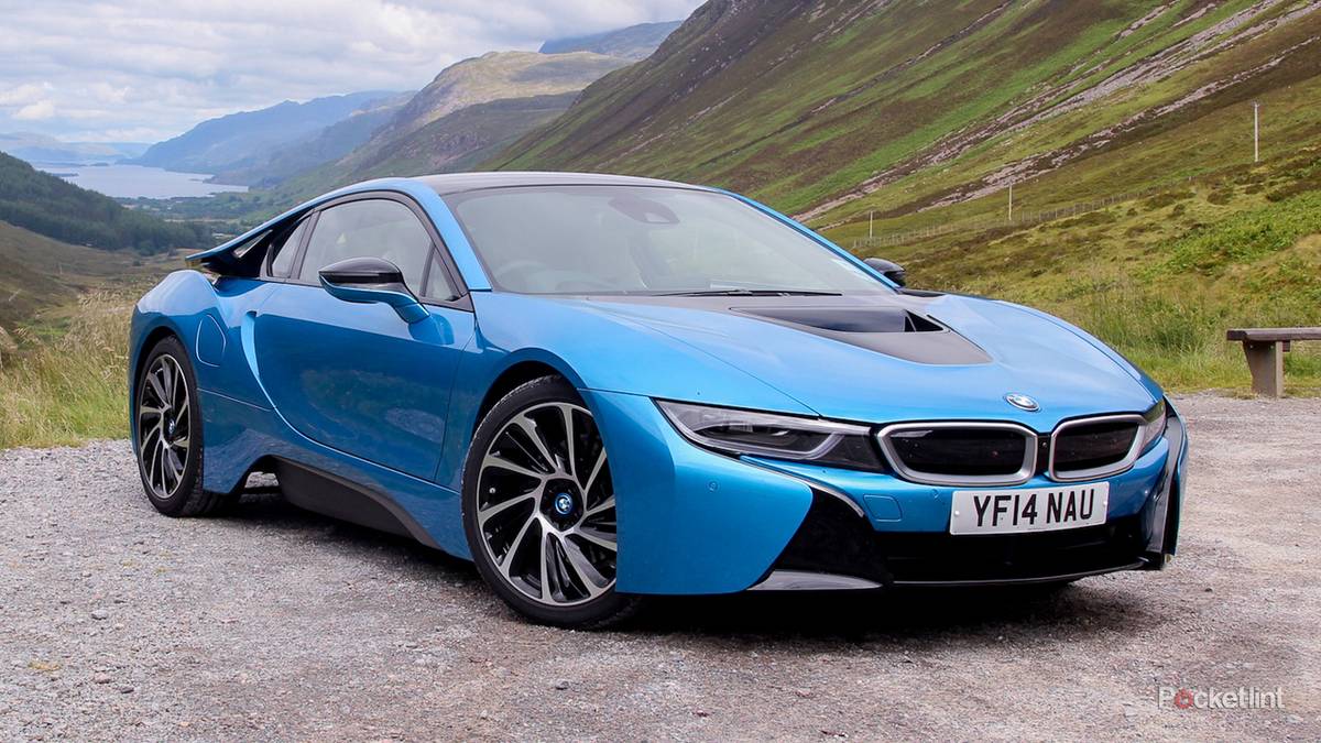 Blue BMW i8 sports car parked on a gravel overlook with green hills and mountains in the background while a lake is visible in the distance.