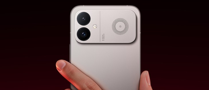Person holds a smartphone by the edges, showing the back with a dual-camera module and a round speaker near the top-right area of the back plate.