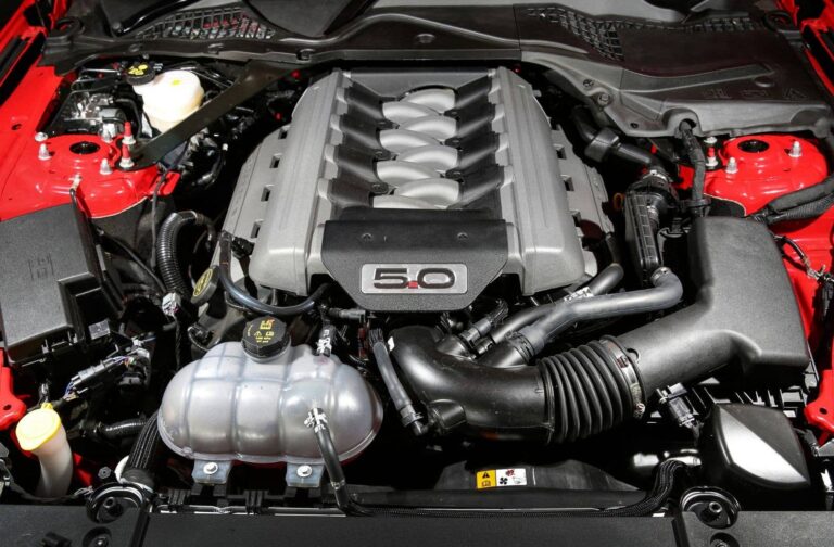 Under-hood view of a car showing a 5.0L V8 engine with a silver intake manifold and surrounding hoses and red chassis.