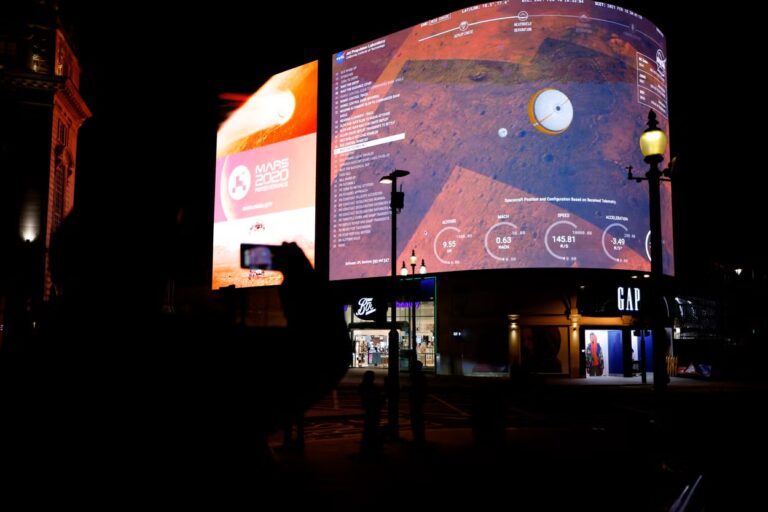 Night scene with two large curved LED screens displaying Mars 2020 Perseverance graphics, silhouettes of people in front of a storefront.