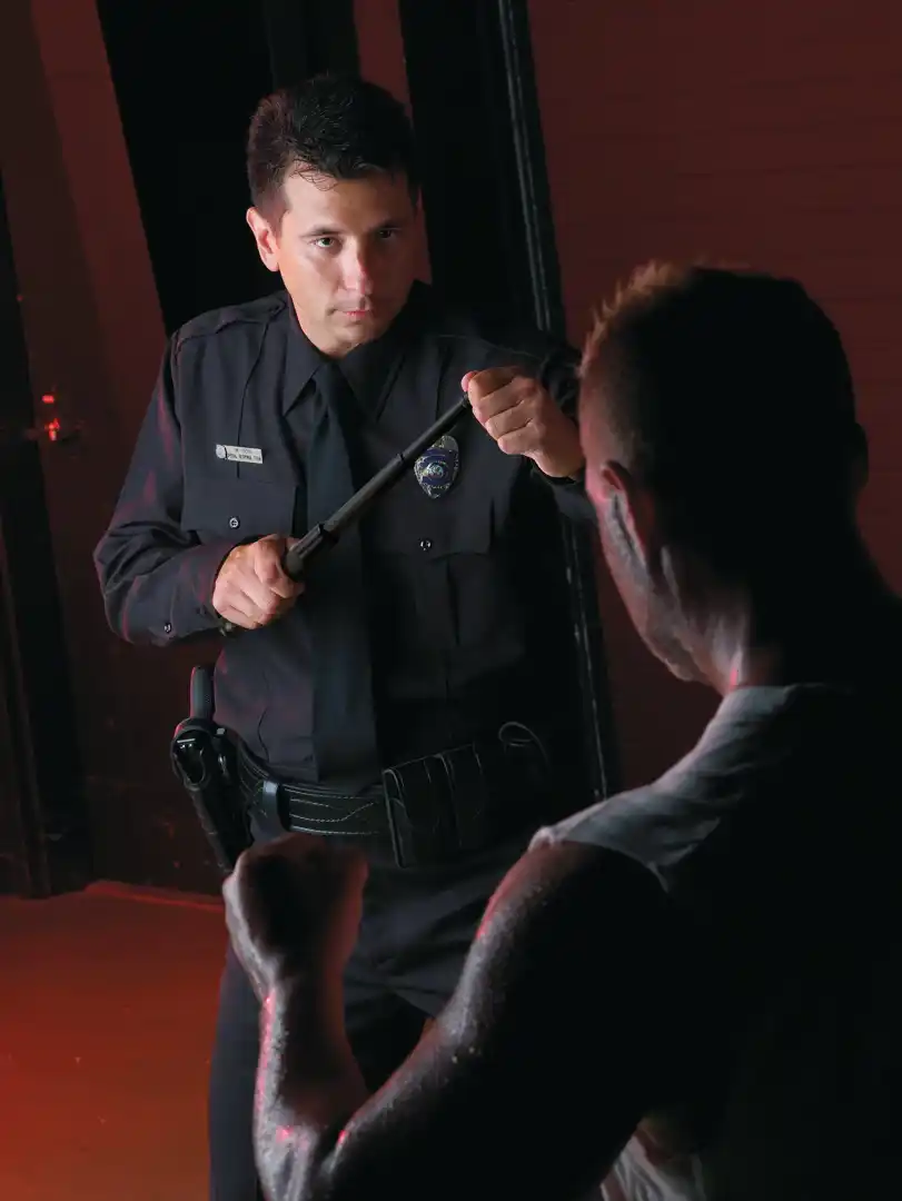 Uniformed officer brandishes a baton at a man facing him in a dim doorway.