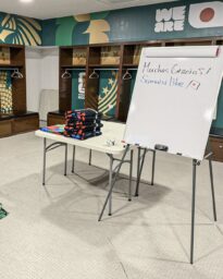 Whiteboard on a stand in a locker room, with folded towels on a table and handwritten message in Spanish thanking Samurai Blue with a small flag icon.