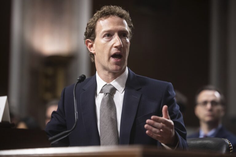 Man in a dark suit and tie speaking at a podium with a microphone in a formal hearing or meeting room.