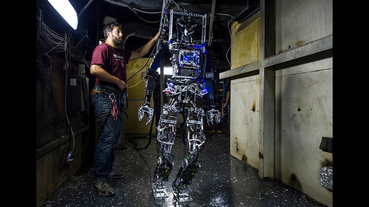 SAFFiR Engineer in a maroon shirt adjusts a tall metallic humanoid robot exoskeleton in a dim workshop.