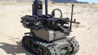 Unmanned ground vehicle (military robot) with mounted machine gun and multiple barrel tubes in a desert setting. It appears to be a remote-weapon platform on tracked treads.