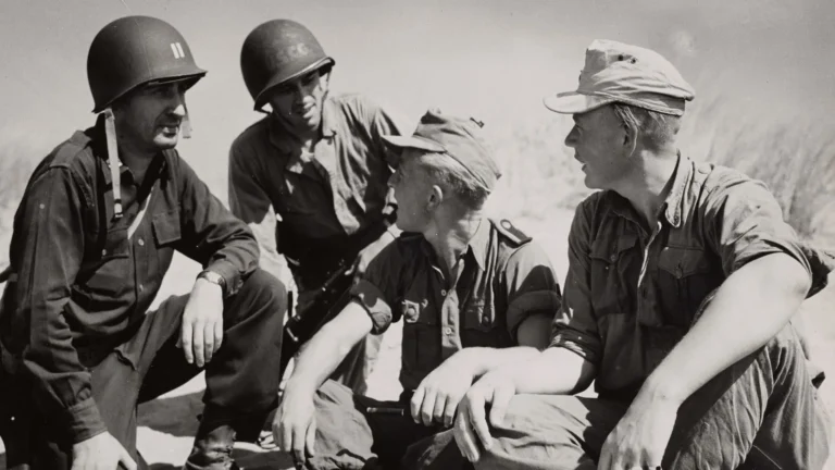 Group of four soldiers in field uniforms sit outdoors, chatting with relaxed postures and helmets on their heads.