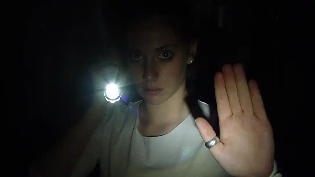 Woman in a dark space shines a bright flashlight toward the camera, palm raised in a stop gesture.
