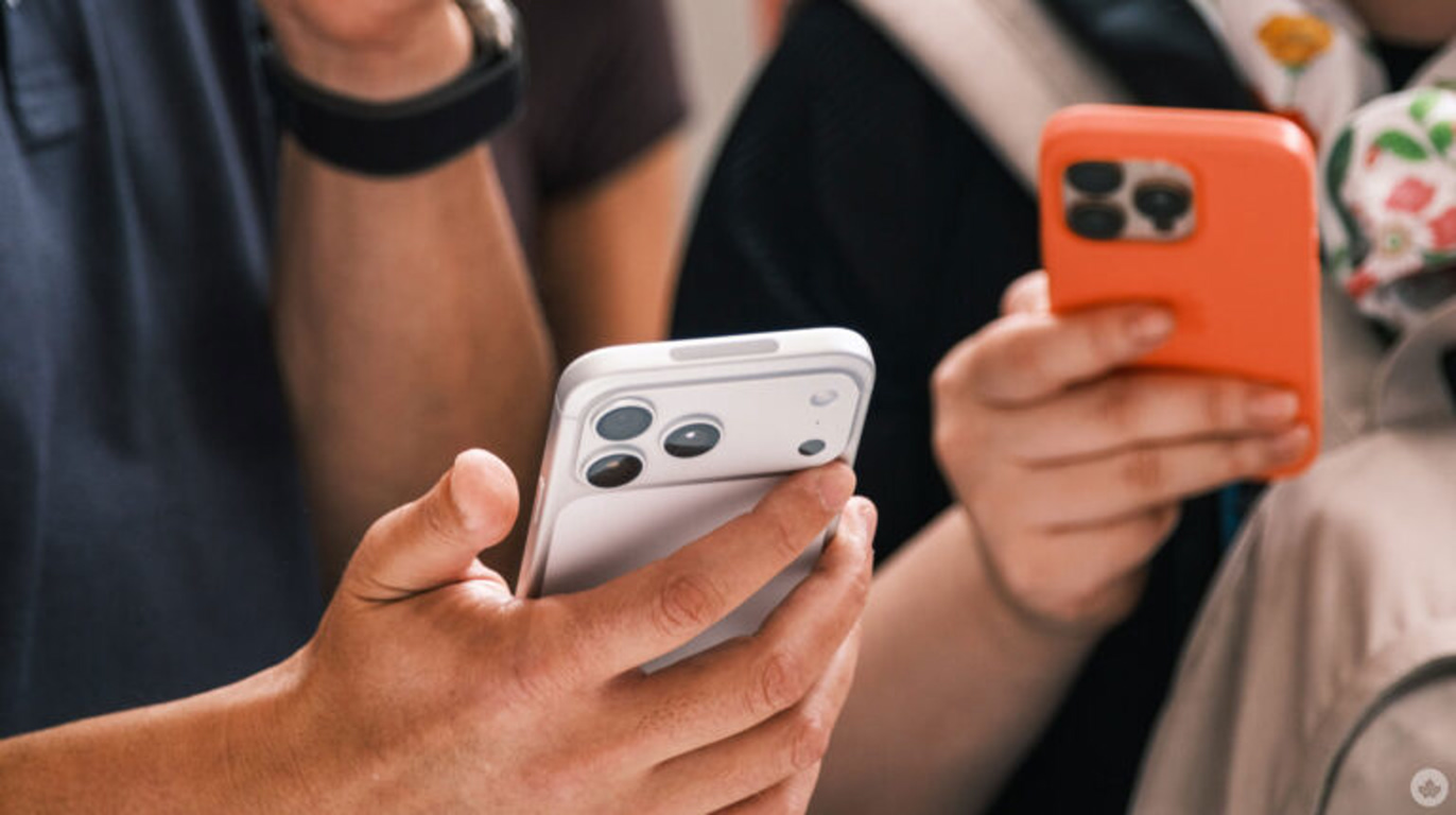 Two people hold smartphones; a white iPhone in the foreground and an orange-case phone in the background.