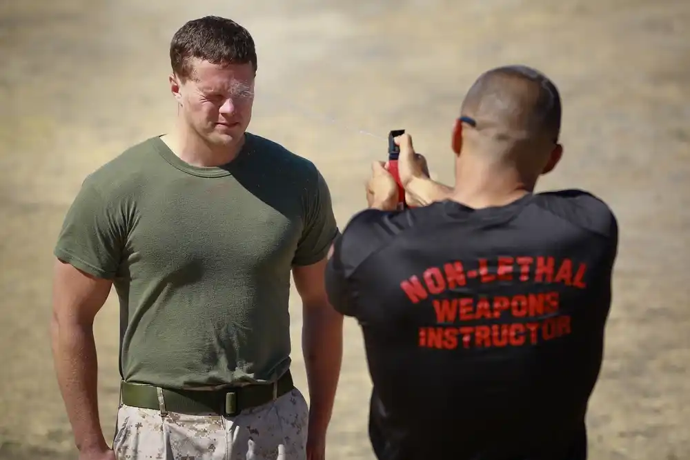 A muscular man in a green t-shirt squints as another man in a black shirt labeled 'Non-Lethal Weapons Instructor' aims a device at him from behind.