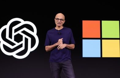 Speaker in a dark stage, wearing a purple shirt, gesturing while presenting; large abstract logo on the left and a four-colored square grid on the right as backdrop.