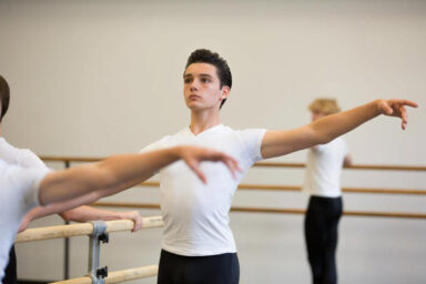 Young male ballet dancer in a white T-shirt stretches at a barre with arms extended in a dance studio.