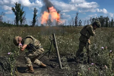 RUSSIA Two soldiers in camouflage gear crouch in a grassy field as a mortar fires, flames and black smoke plume rising in the distance.