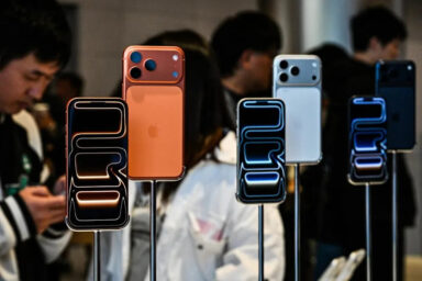 Multiple iPhones on display stands at a tech event, in orange, white, and blue colors with blurred crowd in the background