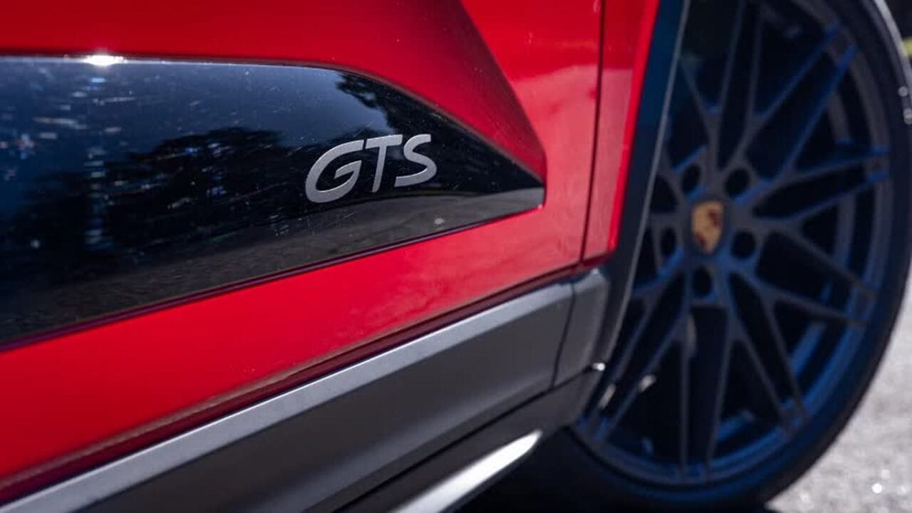 Close-up of a red sports car with a black side panel badge reading 'GTS' and a blue multi-spoke wheel in the background (Porsche).
