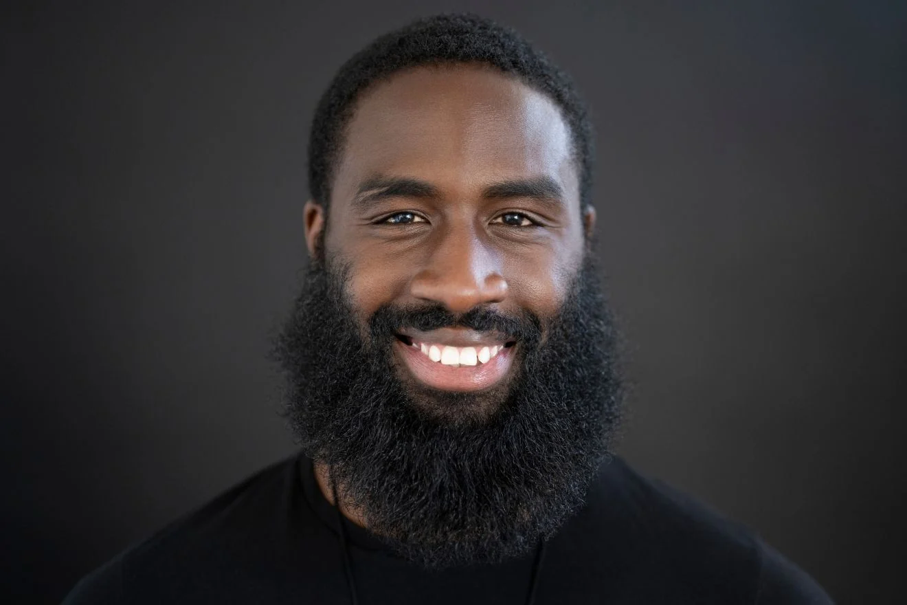 Close-up portrait of a smiling man with a thick beard and short hair against a dark background.