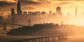 City skyline at sunrise with a curved suspension bridge spanning the water and a hazy orange glow.