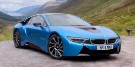 Blue BMW i8 sports car parked on a gravel overlook with green hills and mountains in the background while a lake is visible in the distance.