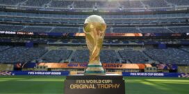 Golden FIFA World Cup Original Trophy on a pedestal in an empty stadium, banners read 'FIFA World Cup 2026'.