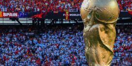 Golden FIFA World Cup trophy in the foreground, with a packed stadium of red and white-clad fans behind; a Spain banner visible in the stands.