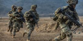 Group of soldiers in camouflage sprinting across a dry field, each carrying rifles and gear with mountains in the background.